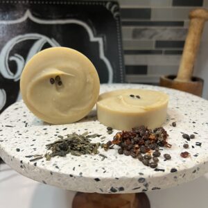 Two light brownish soap rounds on a pedestal stand with green tea leaves and papaya seeds in front.