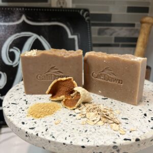 Two soap bars on a pedestal stand with dried orange peels, orange powder and oats in front.