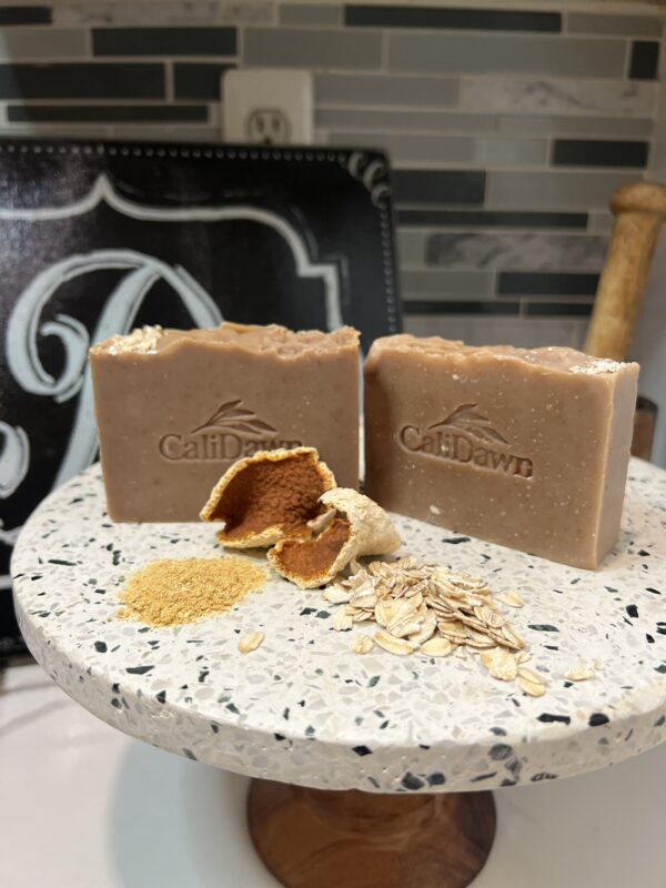 Two soap bars on a pedestal stand with dried orange peels, orange powder and oats in front.