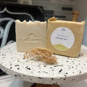 Two soap bars on a pedestal stand, one in a brown packaging with label. Dried sea moss in front of bars.