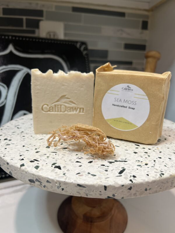 Two soap bars on a pedestal stand, one in a brown packaging with label. Dried sea moss in front of bars.