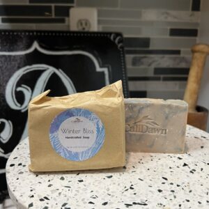 two blue and tan swirl soap bars on a pedestal stand. One in brown package with label.