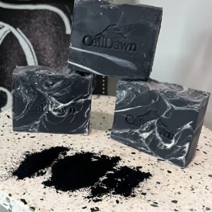 Three Charcoal/Black soap bars with white swirls on a pedestal with powdered charcoal sprinkled in front of bars