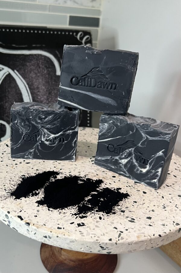 Three Charcoal/Black soap bars with white swirls on a pedestal with powdered charcoal sprinkled in front of bars