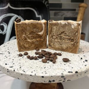 Two coffee and cram colored soaps on a pedestal stand with coffee beans scattered in front.