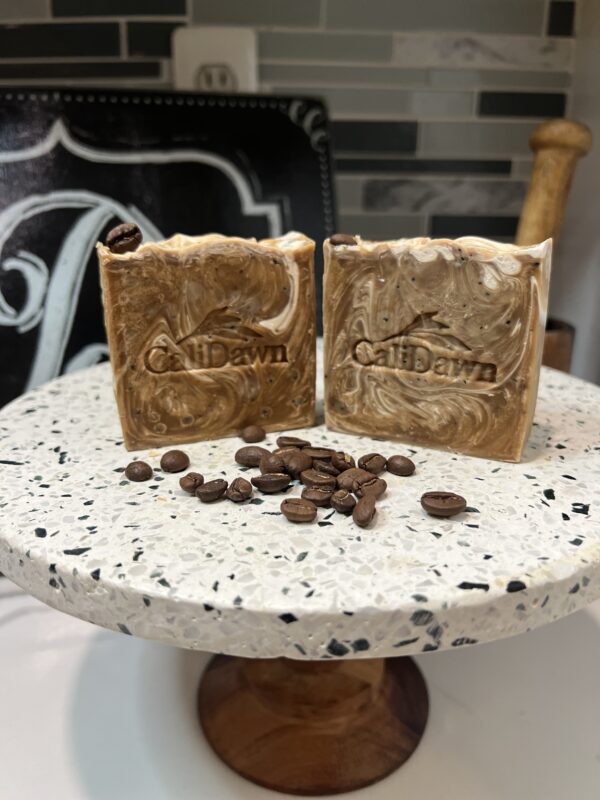 Two coffee and cram colored soaps on a pedestal stand with coffee beans scattered in front.