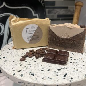 Two coffee and cream colored soaps on pedestal stand with coffee beans and dark chocolate in front.