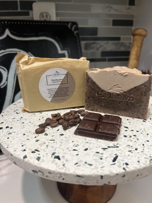 Two coffee and cream colored soaps on pedestal stand with coffee beans and dark chocolate in front.