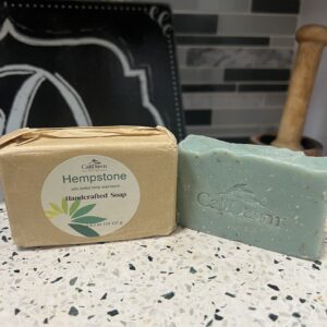 Two soaps sit on pedestal stand. one is unpackaged, light green. The other is in brown package with round label.