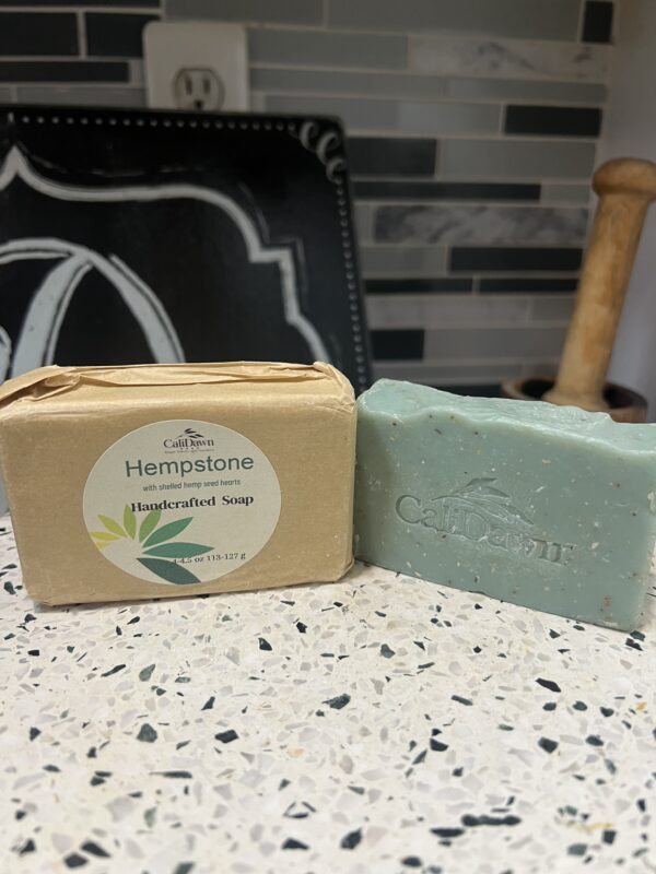 Two soaps sit on pedestal stand. one is unpackaged, light green. The other is in brown package with round label.