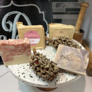 Four Pink and brown soaps on a pedestal stand, 2 are packaged in brown paper with labels. Dried magnolia pods join them.