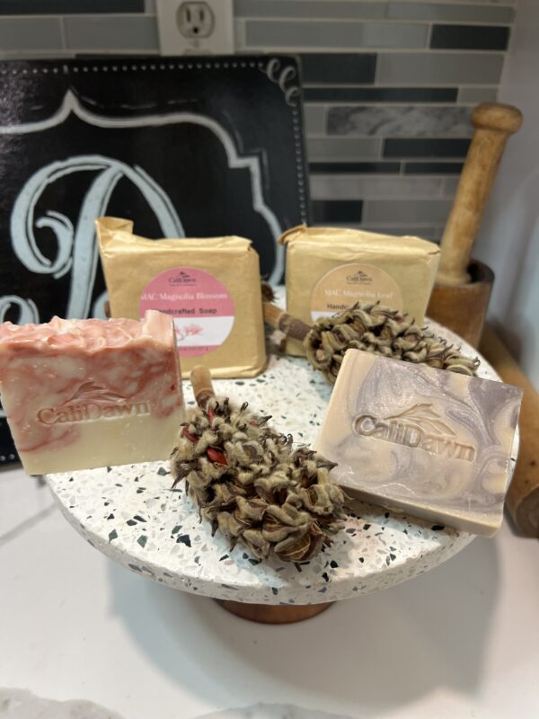 Four Pink and brown soaps on a pedestal stand, 2 are packaged in brown paper with labels. Dried magnolia pods join them.
